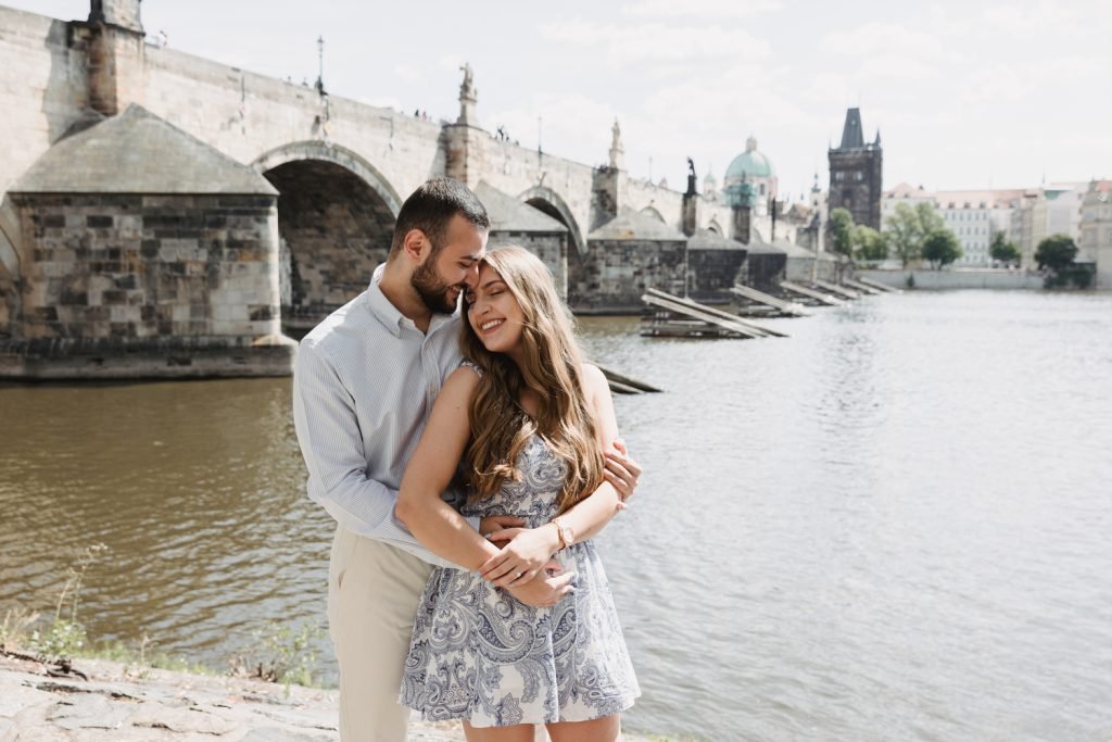 Newly engaged couple by the Vltava with Charles Bridge behind them