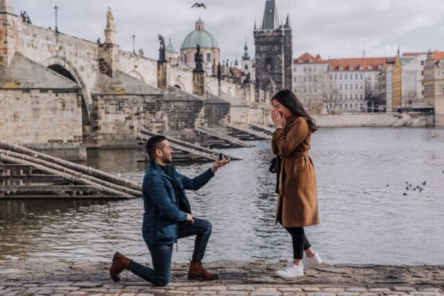 A man kneels to propose to a surprised woman on the Vltava embankment by Charles Bridge in Prague